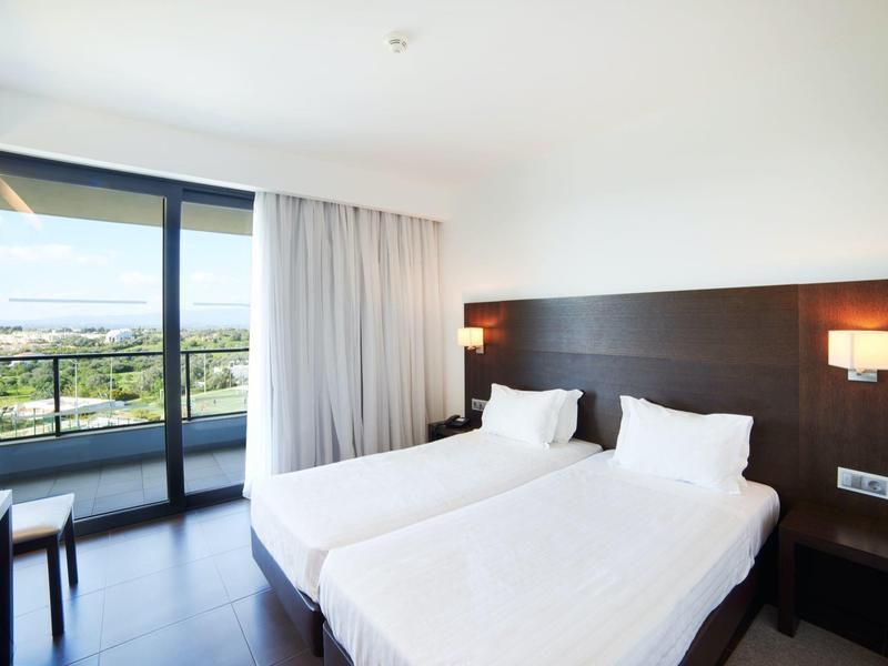 Světý hotelový pokoj se dvěma jednolůžky, tmavým dřevěným nábytkem a balkonem s výhledem na přírodu.