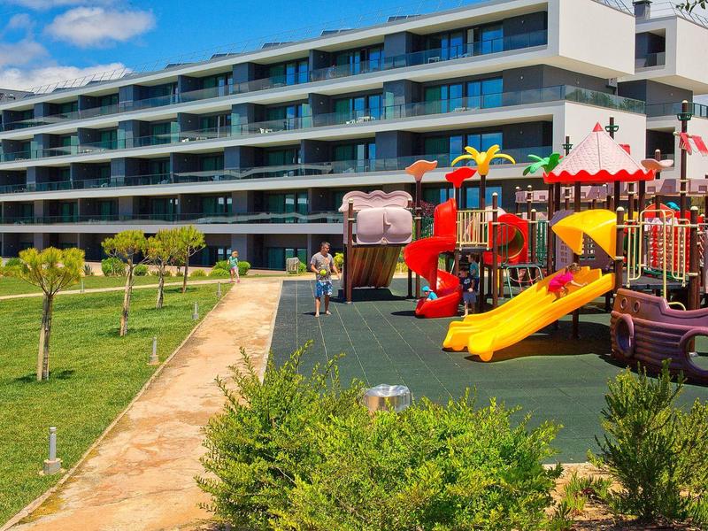 Dětské hřiště se žlutou skluzavkou před moderní hotelovou budovou za slunečného dne