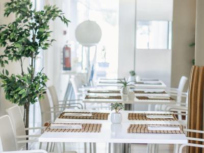 Heller, moderner Gastraum mit weißen Tischen, Stühlen, hängenden Lampen und grünen Pflanzen.