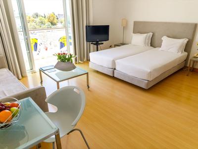 Helles Hotelzimmer mit zwei Betten, Balkon, Fernseher, Tisch und Stühlen, Holzfußboden und moderner Einrichtung.