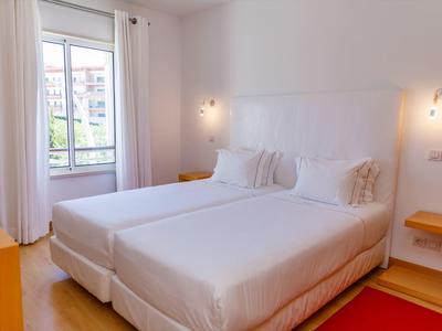 Helles Schlafzimmer mit zwei Einzelbetten, weißen Wänden, Holzfußboden und Fenster mit Vorhängen.