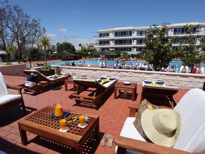 Sonnenterrasse mit weißen Sesseln, Holztischen, Pool und dreistöckigem Gebäude im Hintergrund.