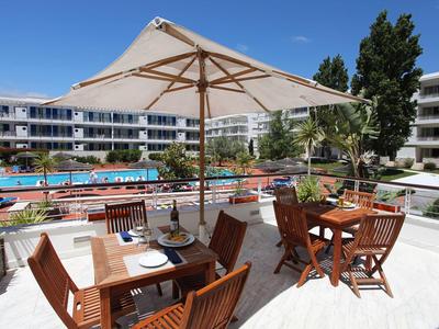 Terrasse mit Holzstühlen, Tischen, Sonnenschirm und Blick auf Pool und angrenzende Gebäude.