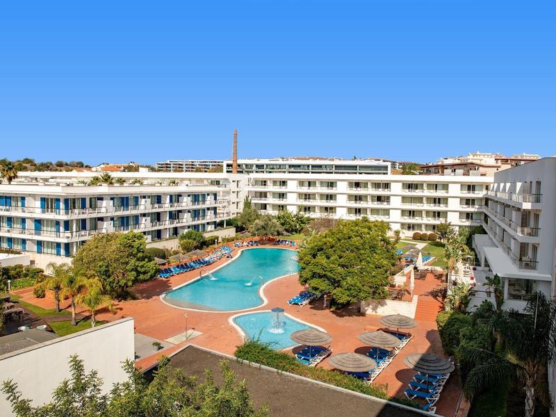 Großes Hotel mit mehreren Etagen und zentralem Swimmingpool, umgeben von Palmen und blauen Himmel.