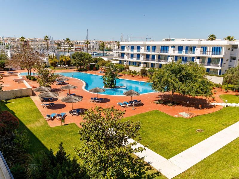 Großer Pool mit Liegen, Palmen und dreistöckigem Gebäude bei sonnigem Himmel.