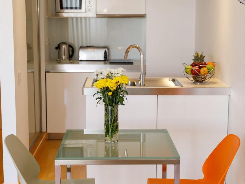 Kleine weiße Küche mit Glastisch, grüner und orangener Stuhl, Blumenstrauß und Obstschale.