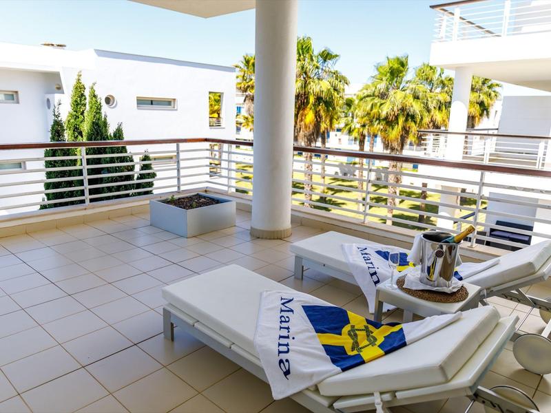 Balkon mit weißen Liegestühlen, Fliesenboden, Palmen und moderner Architektur im Hintergrund.
