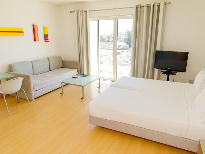 Hell eingerichtetes Hotelzimmer mit Doppelbett, Sofa, Tisch, Fernseher und Balkonfenster mit Vorhängen.