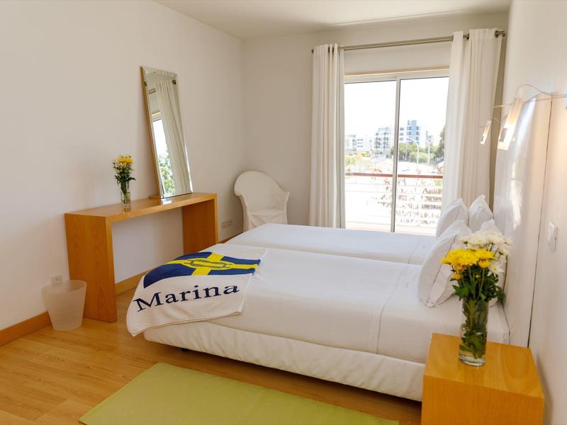 Helles Schlafzimmer mit zwei Betten, Holzfußboden, Spiegel, weißen Vorhängen und Blumen auf Nachttisch.