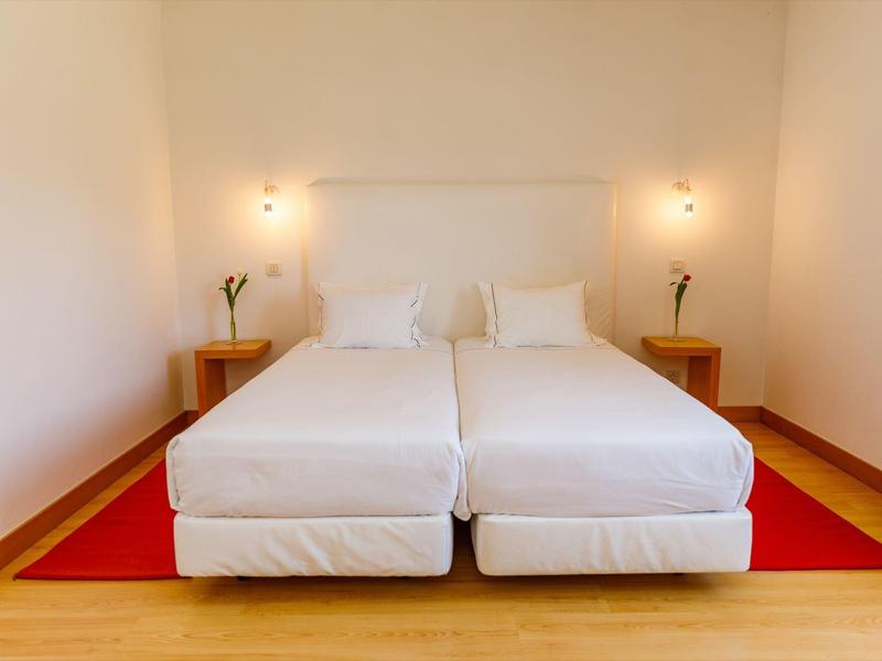 Zwei Einzelbetten mit weißer Bettwäsche auf rotem Teppich in schlicht eingerichteter, heller Ecke.