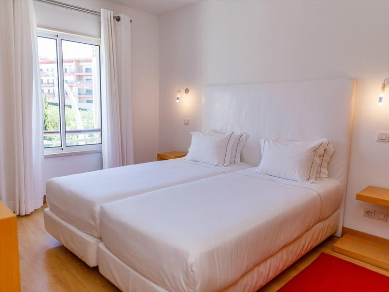 Helles Schlafzimmer mit zwei Einzelbetten, weißen Wänden, Holzfußboden und Fenster mit Vorhängen.