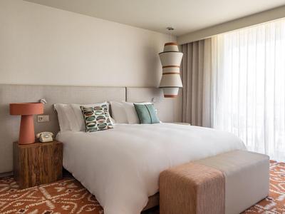 Chambre d'hôtel lumineuse avec grand lit, porte-fenêtre, tapis et décoration moderne.