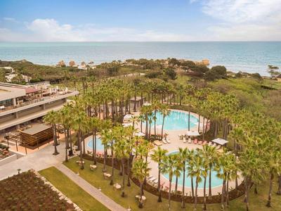 Complexe hôtelier avec deux piscines et palmiers près de la plage sous un ciel nuageux.