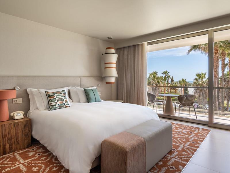Chambre d'hôtel moderne avec lit double, balcon et vue sur des palmiers sous un ciel dégagé.