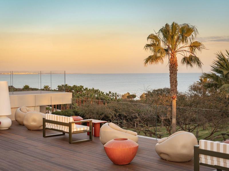 Terrasse avec mobilier moderne, palmiers et vue sur la mer au coucher du soleil.