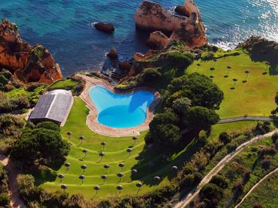 Vista aerea di un hotel con piscina, circondato da prato verde e rocce vicino al mare.