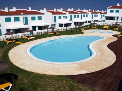 Una piscina curva circondata da case a schiera bianche con tetti rossi e sedie a sdraio gialle.