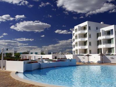 Edificio di appartamenti bianco accanto a una piscina limpida sotto un cielo blu con nuvole.