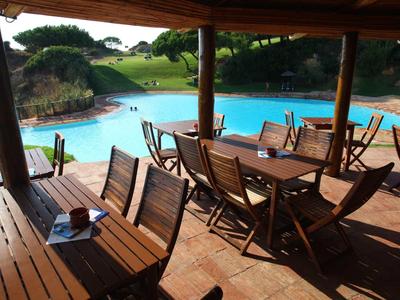 Terrazza coperta con tavoli e sedie di legno accanto a una grande piscina su un prato verde.