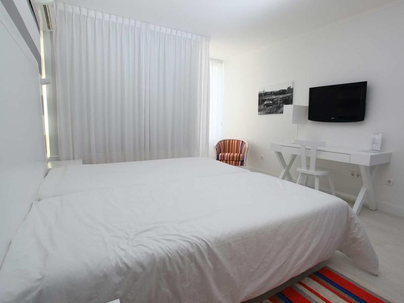 Camera d'hotel luminosa con letto bianco, scrivania, TV e finestra con tende.