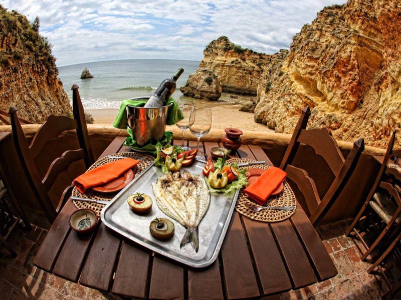 Tavolo con vino, bicchieri e cibo su terrazza con vista su baia rocciosa e mare.
