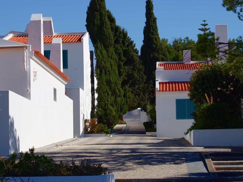 Strada soleggiata con case bianche e tetti rossi, circondata da alberi verdi.