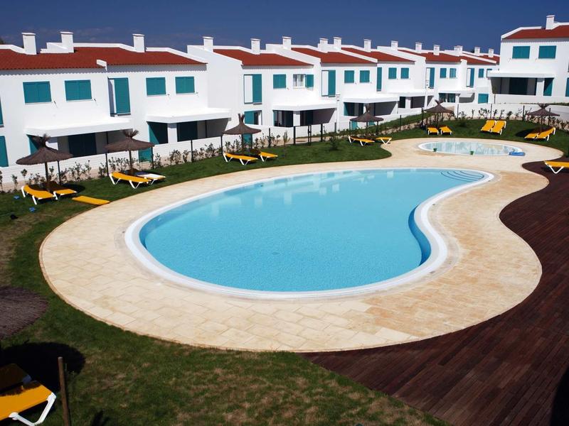 Una piscina curva circondata da case a schiera bianche con tetti rossi e sedie a sdraio gialle.