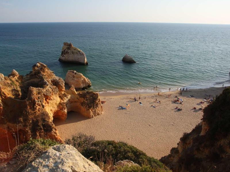 Spiaggia con rocce in acqua e persone sulla sabbia, circondata da scogliere e mare calmo.