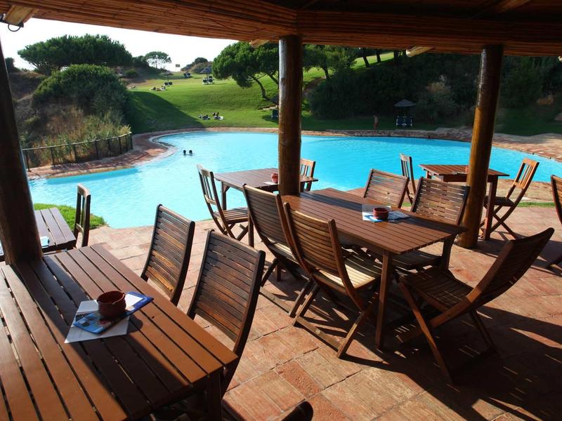Terrazza coperta con tavoli e sedie di legno accanto a una grande piscina su un prato verde.