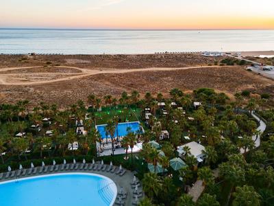 Hotelpoolbereich mit Sonnenliegen und Blick auf Meer und trockene Landschaft im Hintergrund.