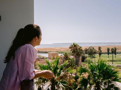 Frau schaut von Hotelbalkon auf Palmen und Meer am sonnigen Tag.