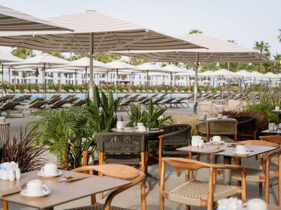 Sonnige Hotelterrasse mit Holztischen, Stühlen und Liegestühlen unter großen Schirmen am Strand.