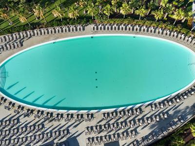 Großer ovaler Pool mit blauen Liegestühlen in einer Hotelanlage, umgeben von Grün.