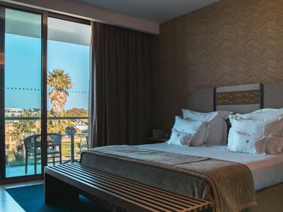 Modernes Hotelzimmer mit Doppelbett und Blick auf Palmen und Terrasse bei Tageslicht.