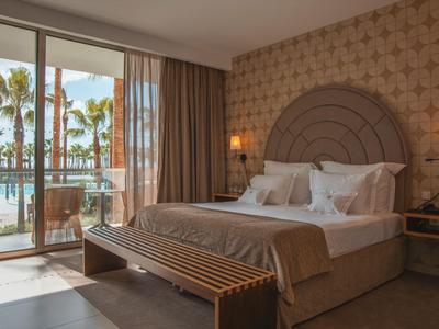 Helles Hotelzimmer mit Doppelbett, Balkon und Blick auf Palmen und Pool.
