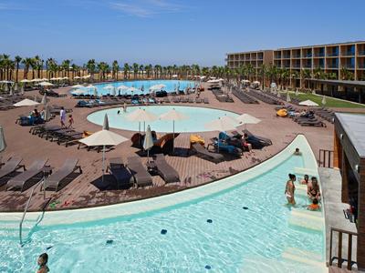 Großer Hotelpool mit Liegestühlen, Sonnenschirmen und Palmen unter blauem Himmel.