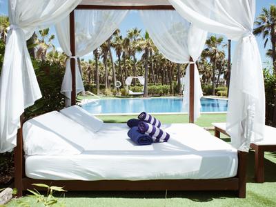 Ein weiß überdachtes Bett mit lila Handtüchern am Pool in tropischer Hotelanlage.