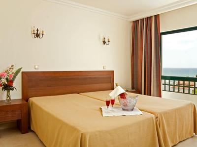 Pokój hotelowy z dwoma łóżkami, drewnianymi meblami, balkonem i widokiem na morze w świetle dziennym.