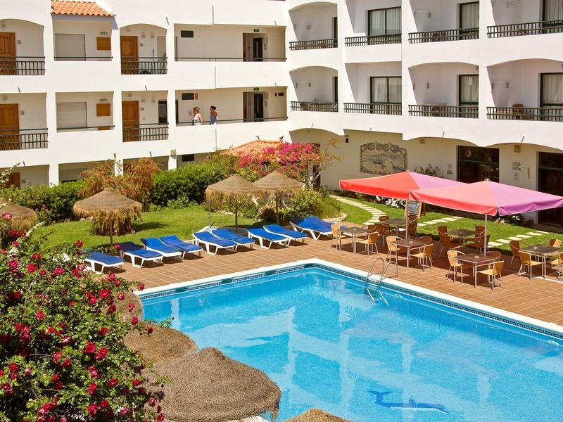 Hotel z basenem na zewnątrz, leżakami, ogrodem i białymi budynkami z balkonami.