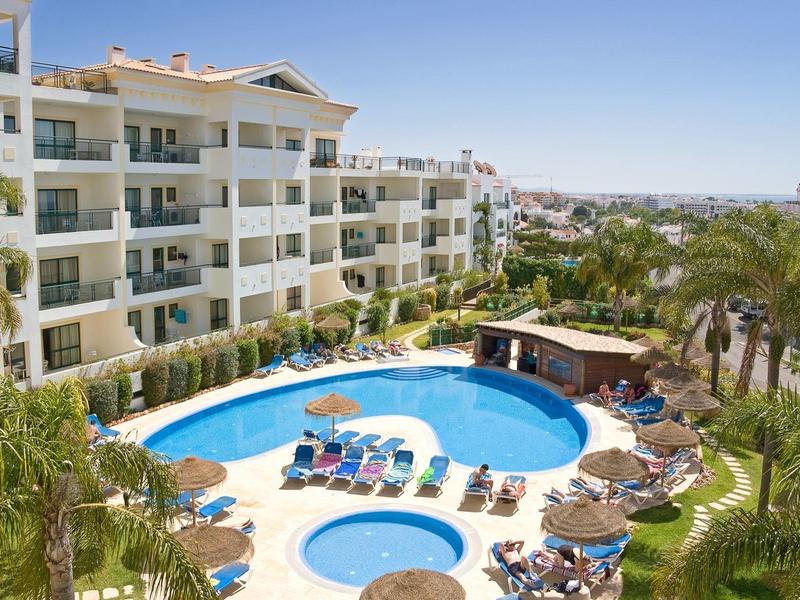 Nowoczesny kompleks apartamentowy z basenem, leżakami i palmami w słonecznym wakacyjnym otoczeniu.