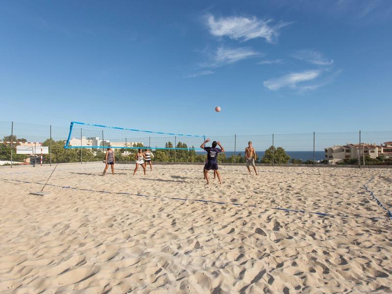Persone che giocano a beach volley su un campo sabbioso con tempo soleggiato e cielo blu.