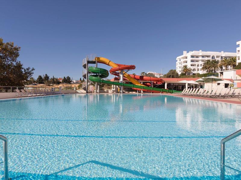 Grande piscina all'aperto con scivoli d'acqua e lettini davanti a edifici hotel bianchi sotto un cielo limpido.