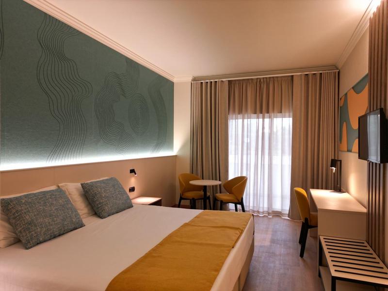 Camera d'hotel moderna con letto matrimoniale, cuscini grigi, accenti gialli e piccolo spazio per sedersi.