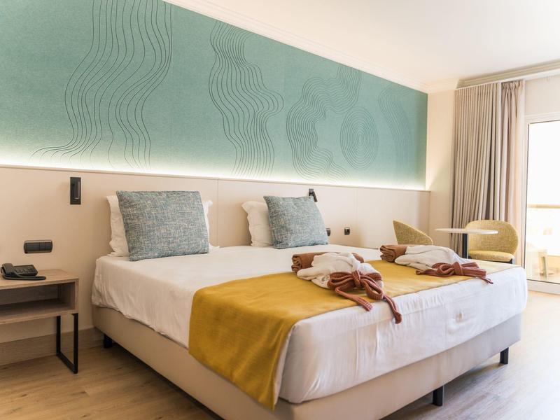 Camera d'hotel moderna con letto matrimoniale, copriletto gialli e balcone con vista sul mare.