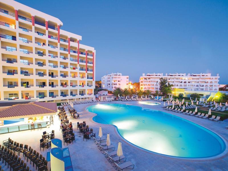 Grande piscina dell'hotel con lettini e edificio alberghiero colorato a più piani al tramonto.
