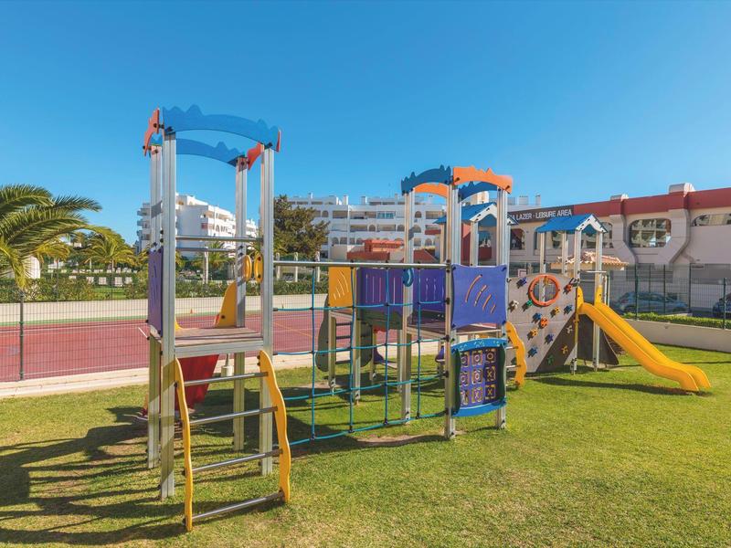 Area giochi per bambini con scivoli e strutture per arrampicarsi su prato davanti a un hotel.