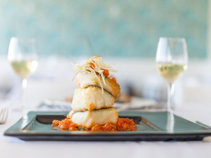Piatto elegante con filetto di pesce e salsa di pomodoro su un piatto nero con due bicchieri di vino bianco sullo sfondo.