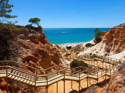 Scale di legno che attraversano rocce rosse verso una spiaggia soleggiata e tranquilla con mare azzurro.