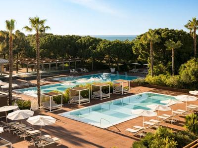 Hotel pool area with loungers, palm trees, and sea view in the background.