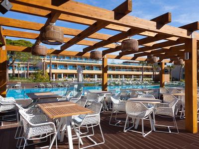 Area pranzo all'aperto con tavoli e sedie sotto una copertura in legno vicino a una piscina dell'hotel.